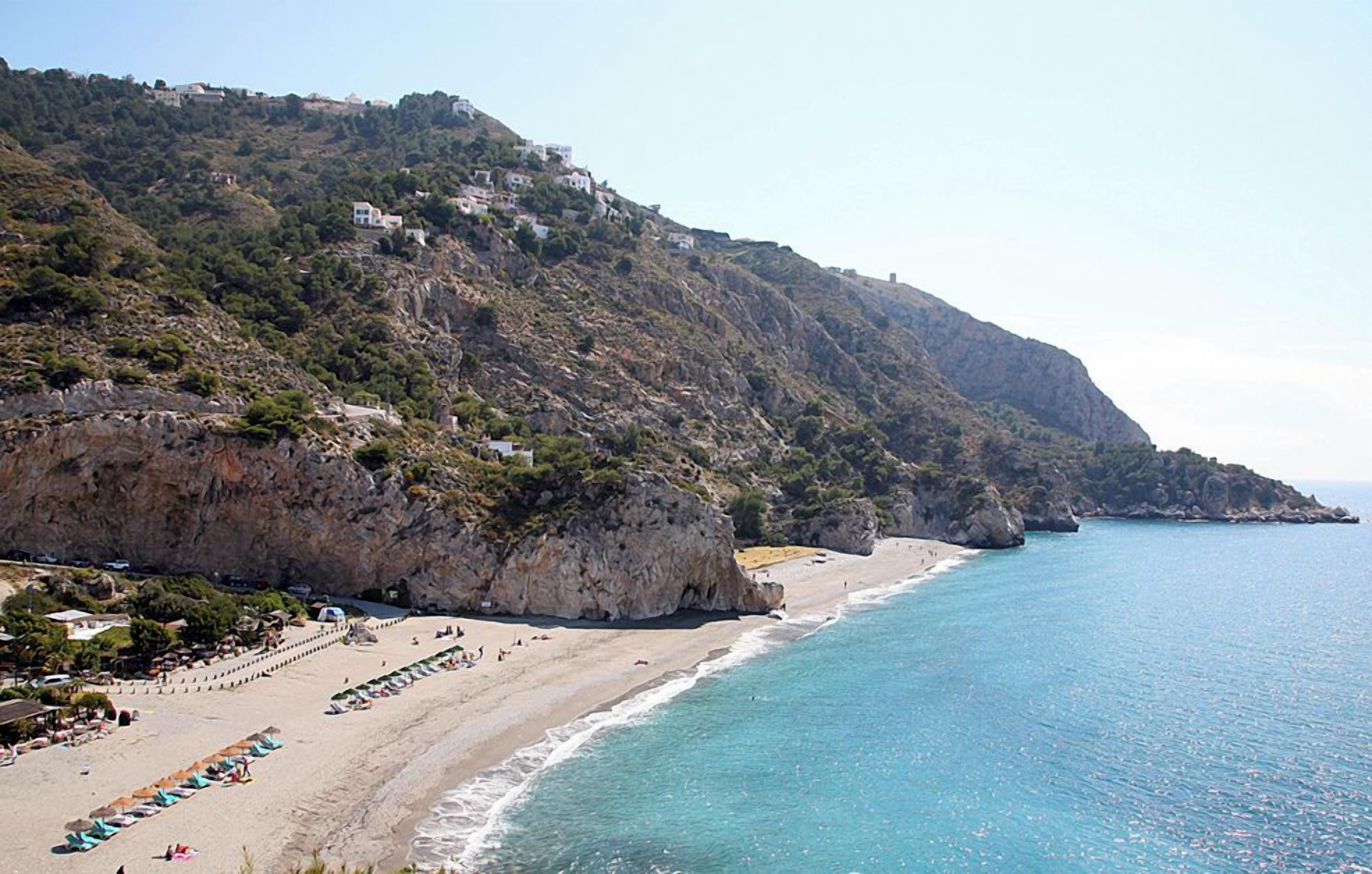 A coastal landscape with a beach, rocky cliffs, and buildings on the hillside.