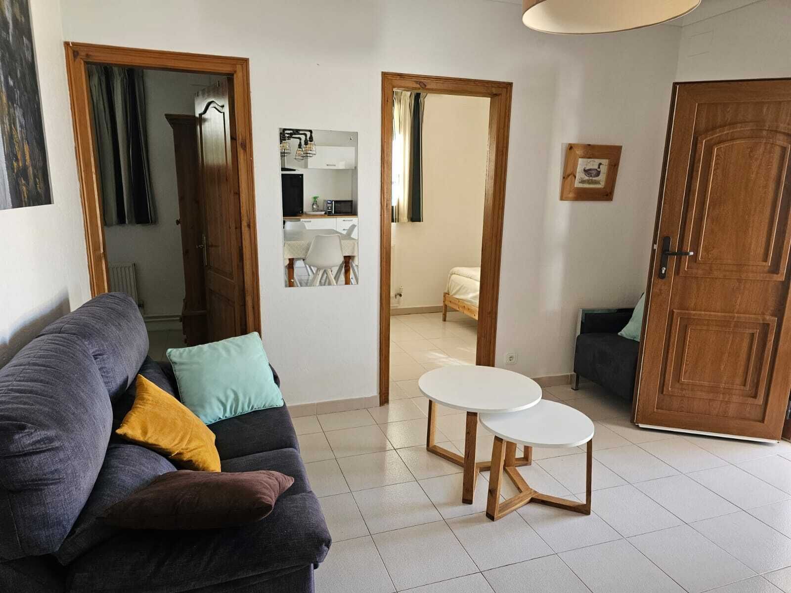 A living room with a sofa, two small tables, a wooden door, and a view into a kitchen and bedroom.