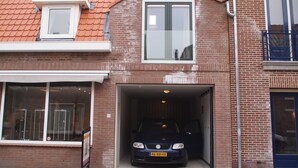 Une voiture est garée à l’intérieur d’un garage attenant à un bâtiment en briques, doté de grandes fenêtres et d’un toit recouvert de tuiles.