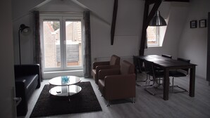 Un salon moderne avec une table ronde en verre, un canapé de couleur sombre, une table à manger avec des chaises et une grande fenêtre donnant sur un bâtiment en briques.