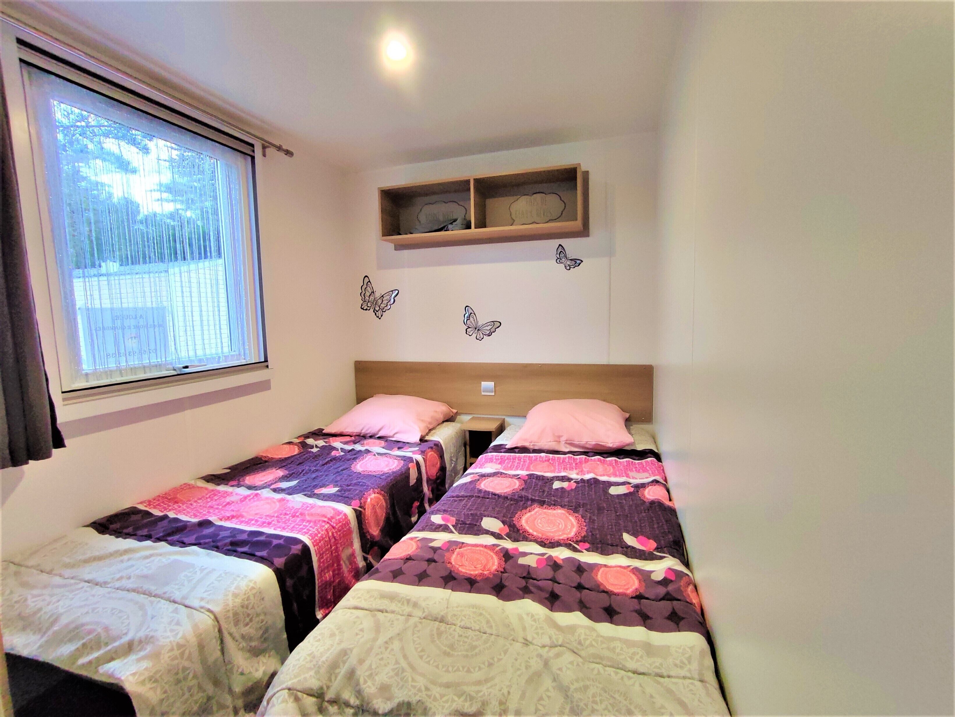 A bedroom with two single beds, a window with blinds, a shelf with decorative items, and butterfly wall decals.