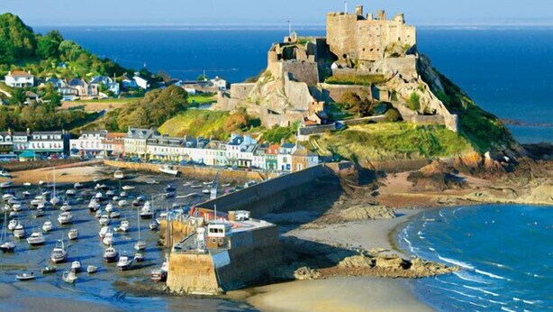 Uma vila costeira com um castelo histórico numa falésia, um porto com barcos e edifícios coloridos ao longo da costa.