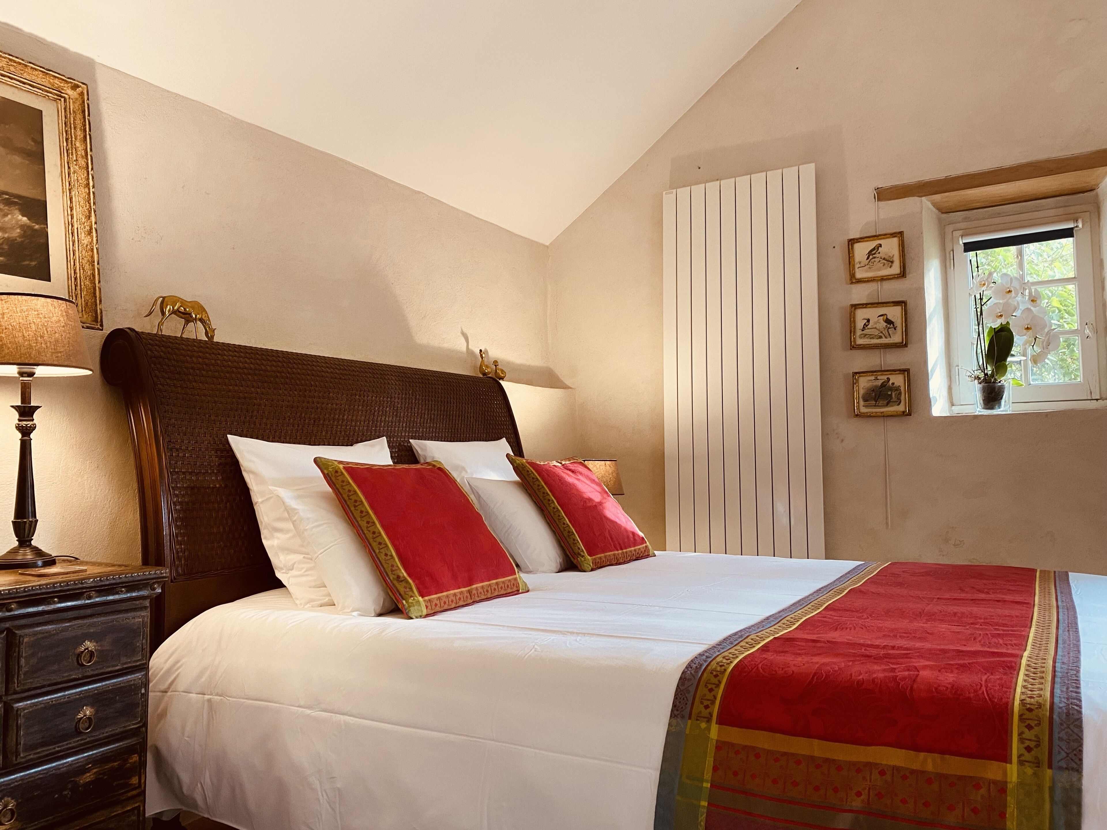 A bedroom with a large bed, a nightstand with a lamp, a window with a view of greenery, and a wall with framed pictures.