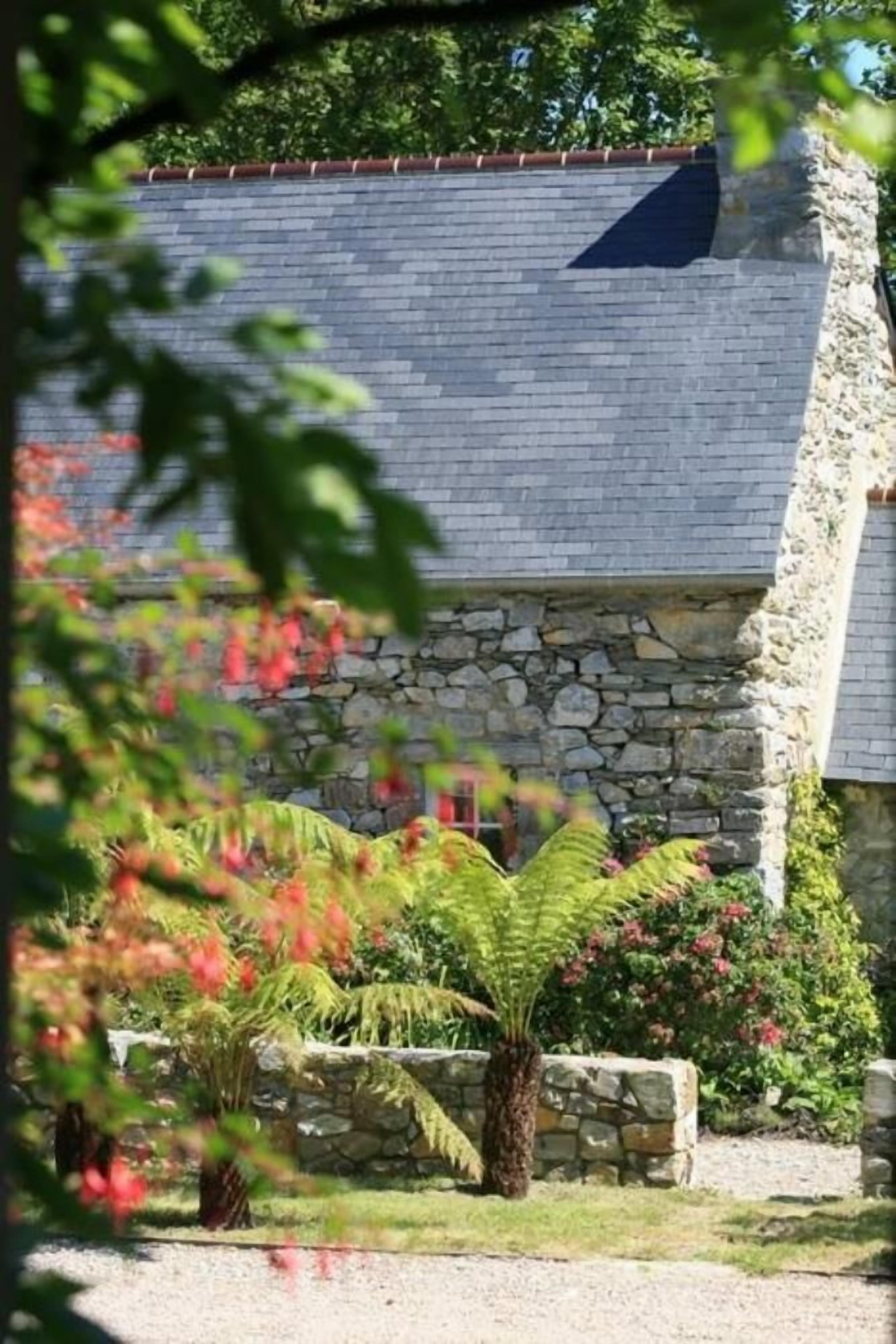 Un bâtiment en pierre doté d’un toit en pente, entouré d’un jardin où poussent différentes plantes et une fougère.