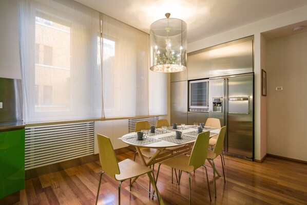 A modern kitchen with a stainless steel refrigerator, a dining table with chairs, and a large window with sheer curtains.
