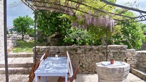 Un área de comedor al aire libre con una mesa y sillas, un muro de piedra y una pérgola adornada con flores moradas colgantes.