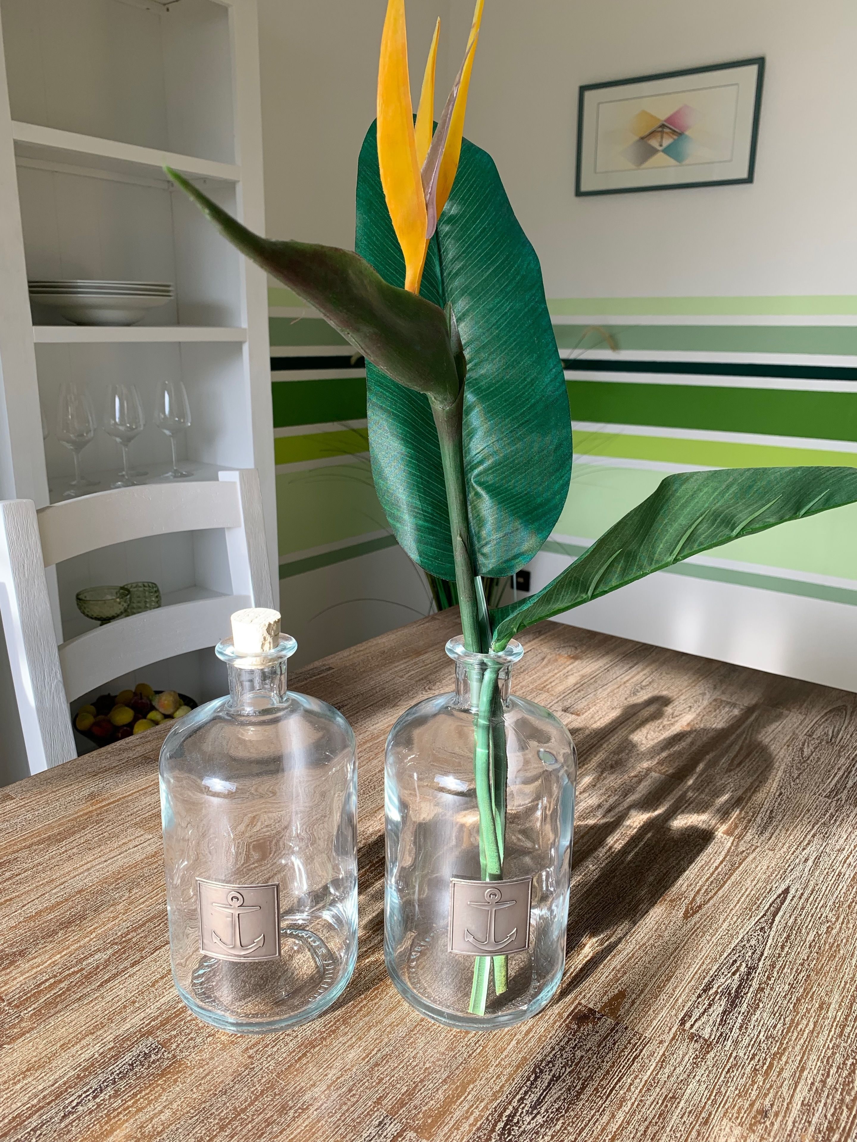 Zwei Glasflaschen mit Anker-Emblemen auf einem Holztisch, eine grüne Pflanze mit gelb-grünen Blättern und im Hintergrund ein weißes Regal mit Weingläsern und Schalen.
