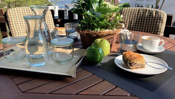 Una mesa dispuesta con una bandeja de frascos de vidrio, una manzana verde, un vaso de agua, una taza de café y una porción de pastel sobre un mantel a cuadros.