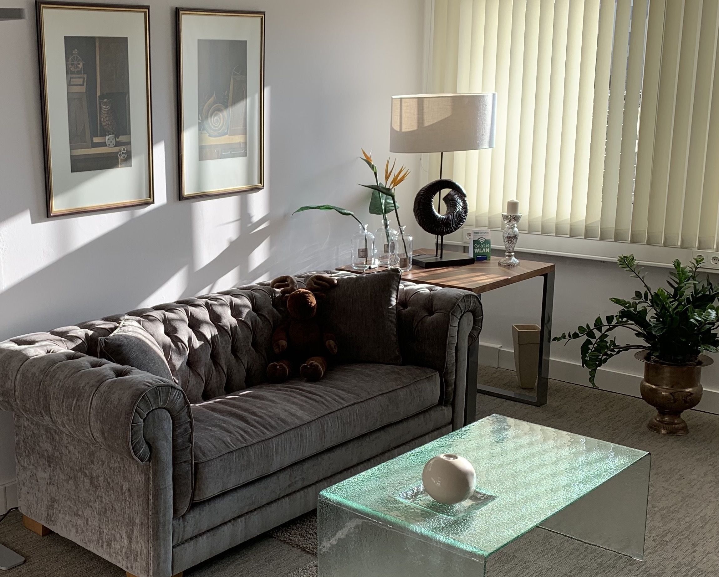 Un salon avec un canapé gris, une table basse en verre, des œuvres d’art encadrées accrochées au mur, une lampe et une plante en pot.