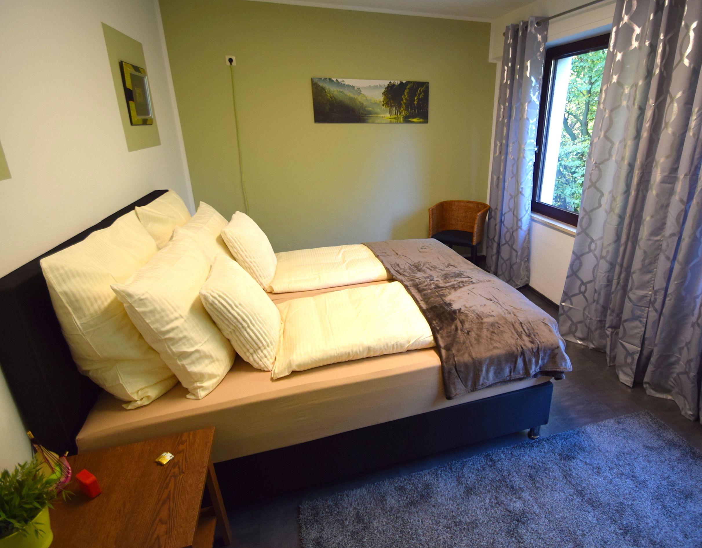 A single bed with a wooden frame and a headboard, a small bedside table with a plant, a window with curtains, and a painting on the wall.