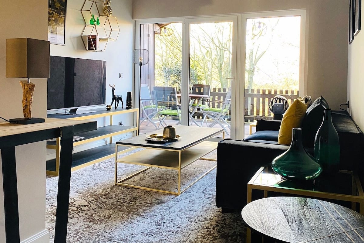 A modern living room with a flat-screen TV, a coffee table, and a sofa. There is a large window providing a view of the outdoors.