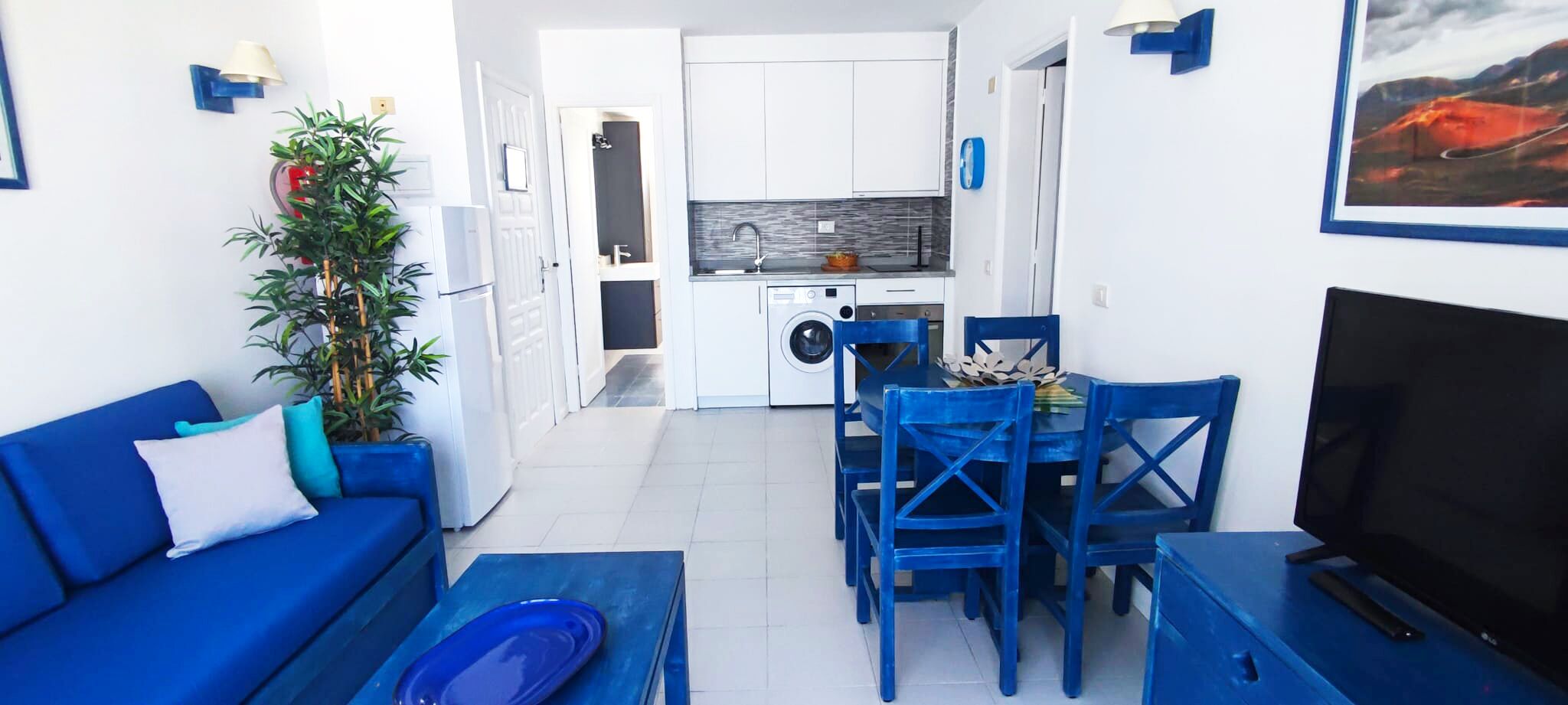 A compact living space with a blue sofa, a dining table with blue chairs, a TV, and a kitchen area with a washing machine.