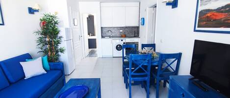 A compact living space with a blue sofa, a dining table with blue chairs, a TV, and a kitchen area with a washing machine.