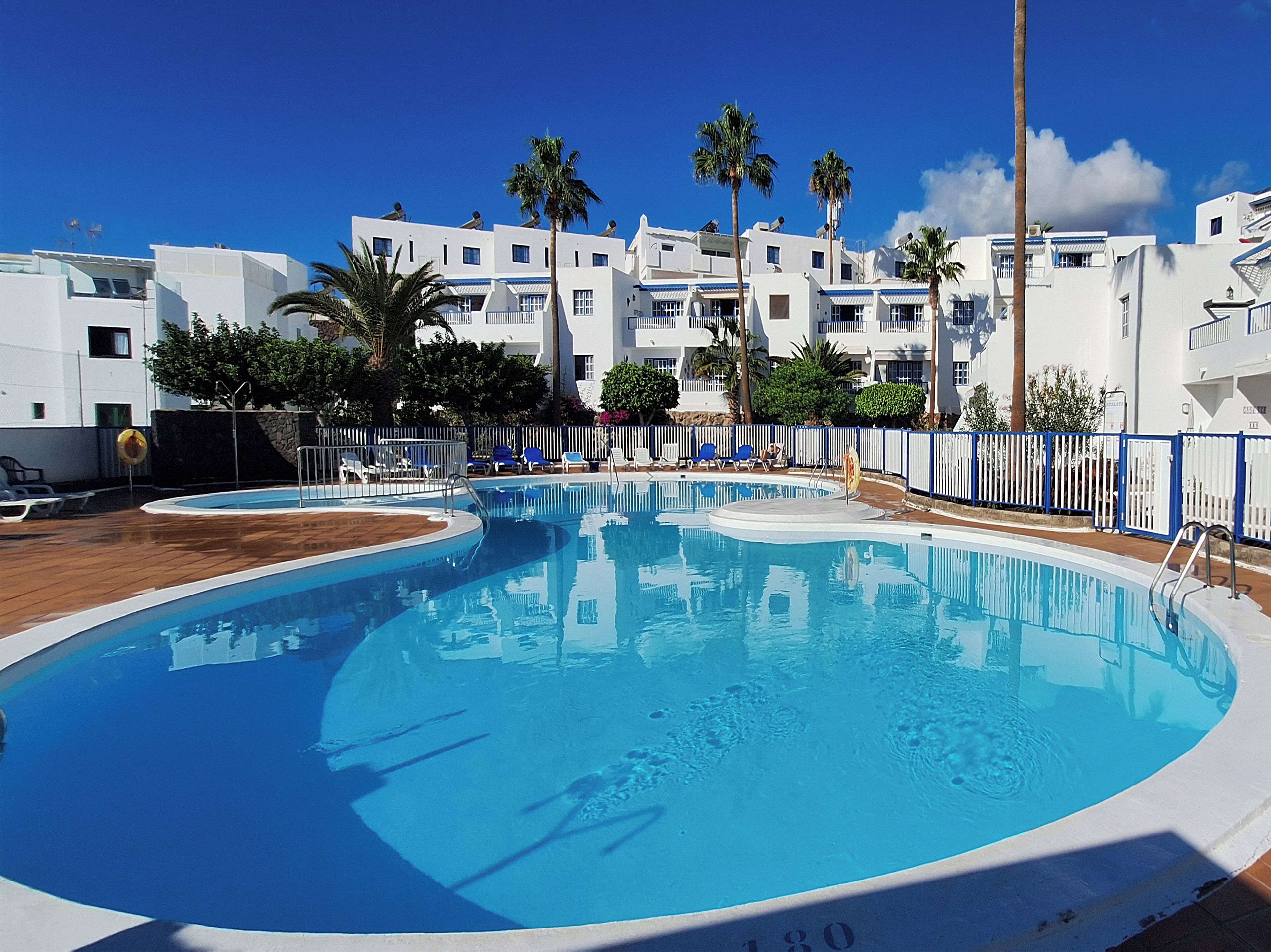 A swimming pool with a wooden deck, surrounded by white buildings with balconies and palm trees.