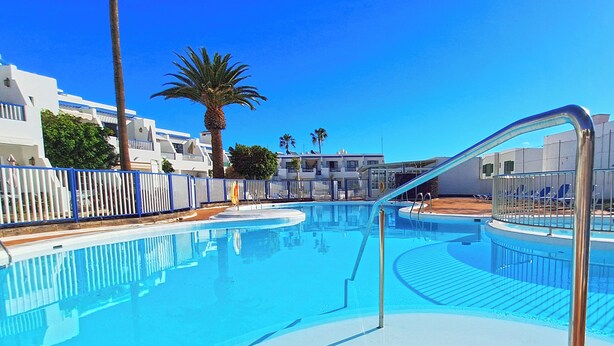 A clear blue swimming pool with a metal ladder, surrounded by white buildings and palm trees. - Sunny and comfy appartment in a residencial development near the sea (Puerto del Carmen, Tias)