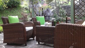 An outdoor seating area with wicker furniture and green cushions, surrounded by lush greenery and a pergola structure. -  Villa with own pool, mountain village with restaurant and shop. North of Lucca (Pieve Fosciana)