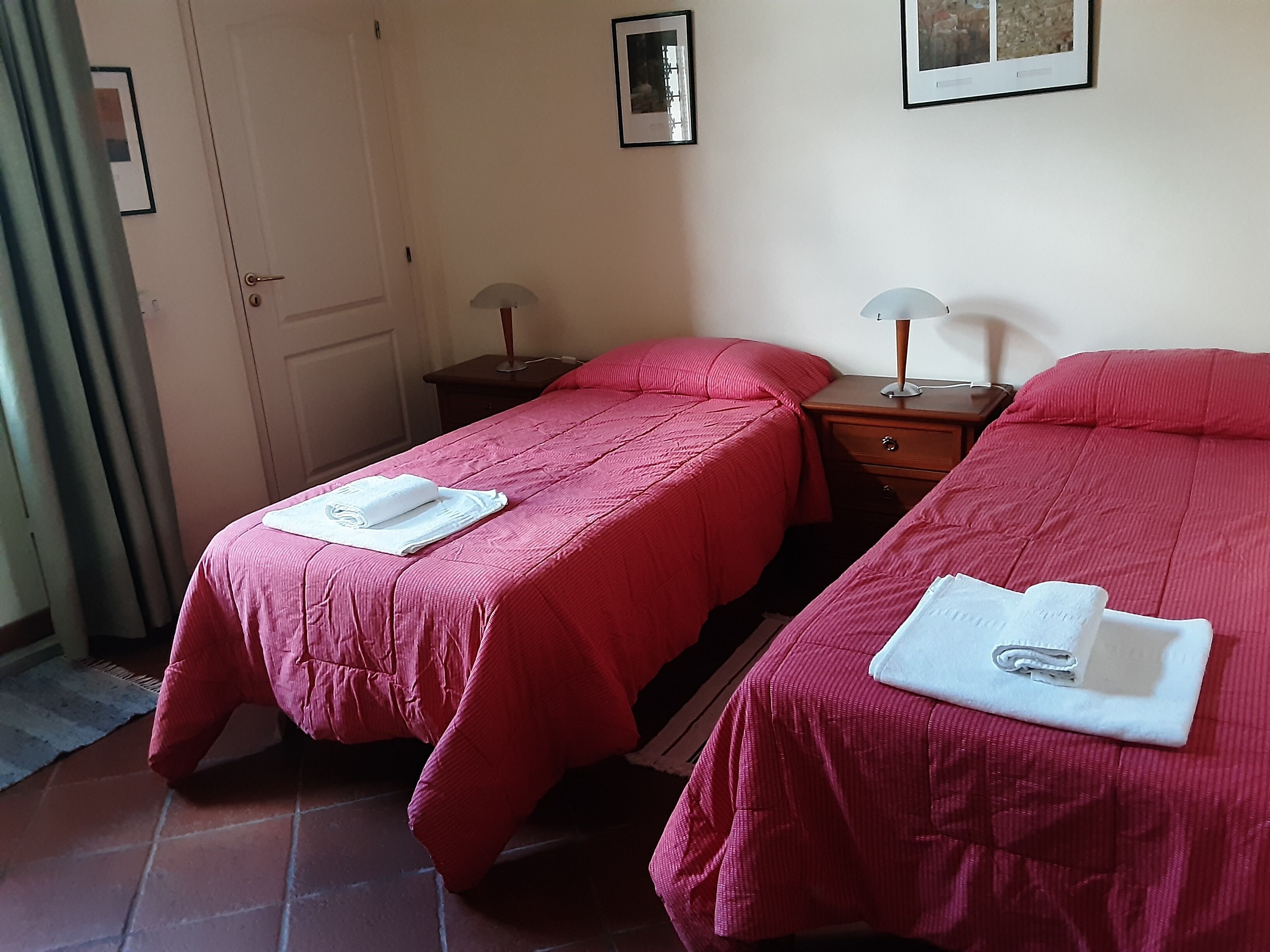 A room with two single beds, each with a red bedspread and a white towel. There are two bedside tables with lamps, a door, and framed pictures on the wall.