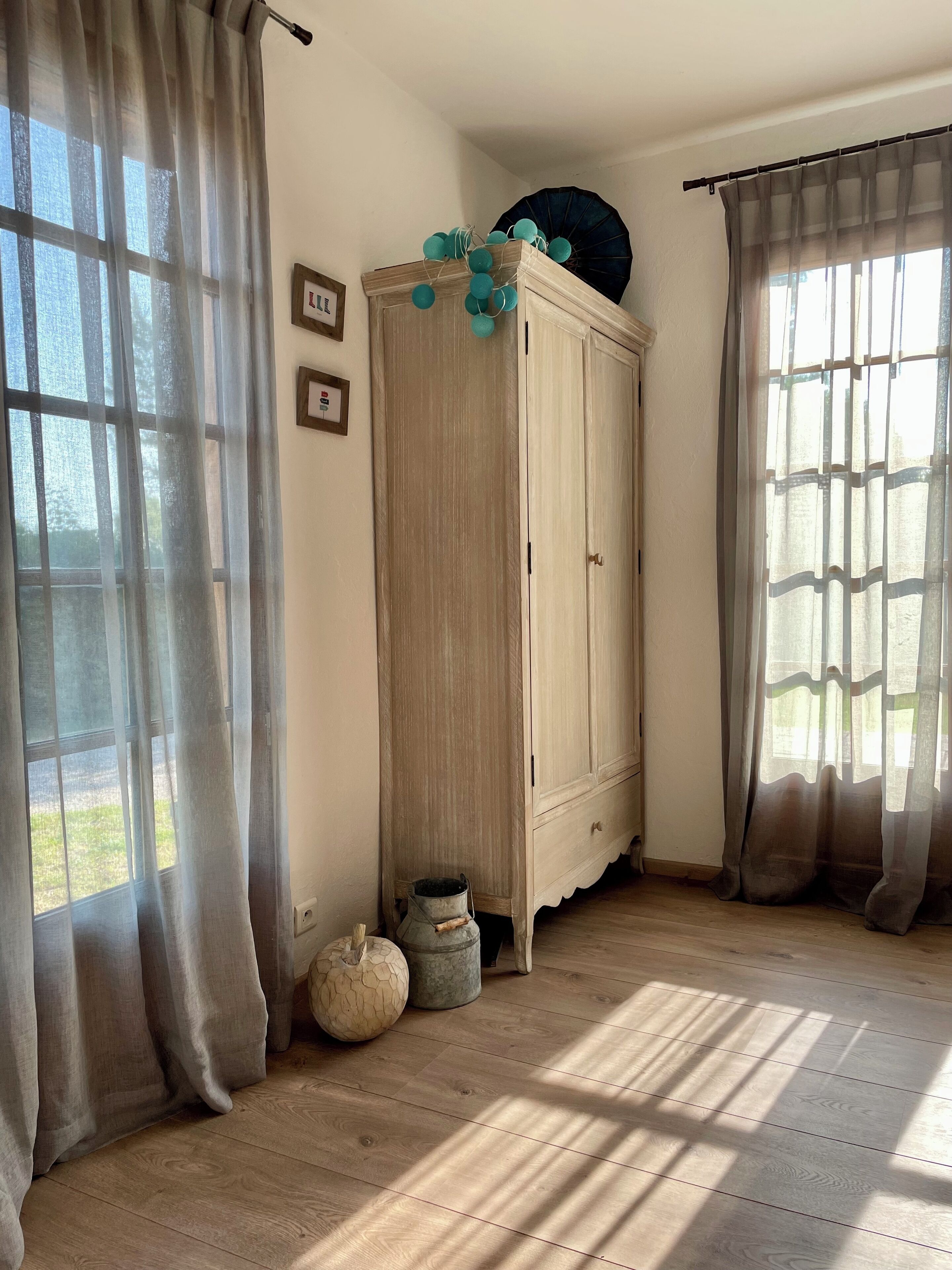 Um quarto com piso de madeira, um guarda-roupa de cor clara e duas janelas com cortinas leves.