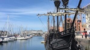 Een haven met een groot zeilschip afgemeerd, omringd door andere boten en gebouwen op de achtergrond.