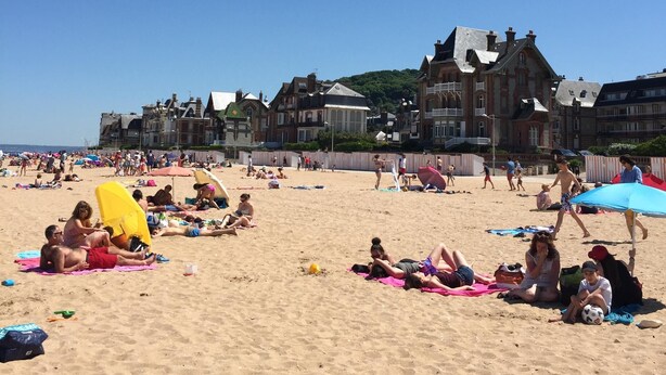 Een strand met mensen die zonnebaden en spelen, een grote gele parasol, en historische gebouwen op de achtergrond.