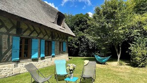 Een traditioneel gebouwd stenen pand met rieten dak en blauwe luiken, omgeven door een gazon en bomen.