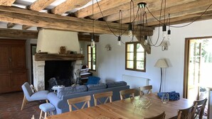 Een rustiek interieur met zichtbare houten balken, een open haard, een eettafel met stoelen en een gezellige zithoek.