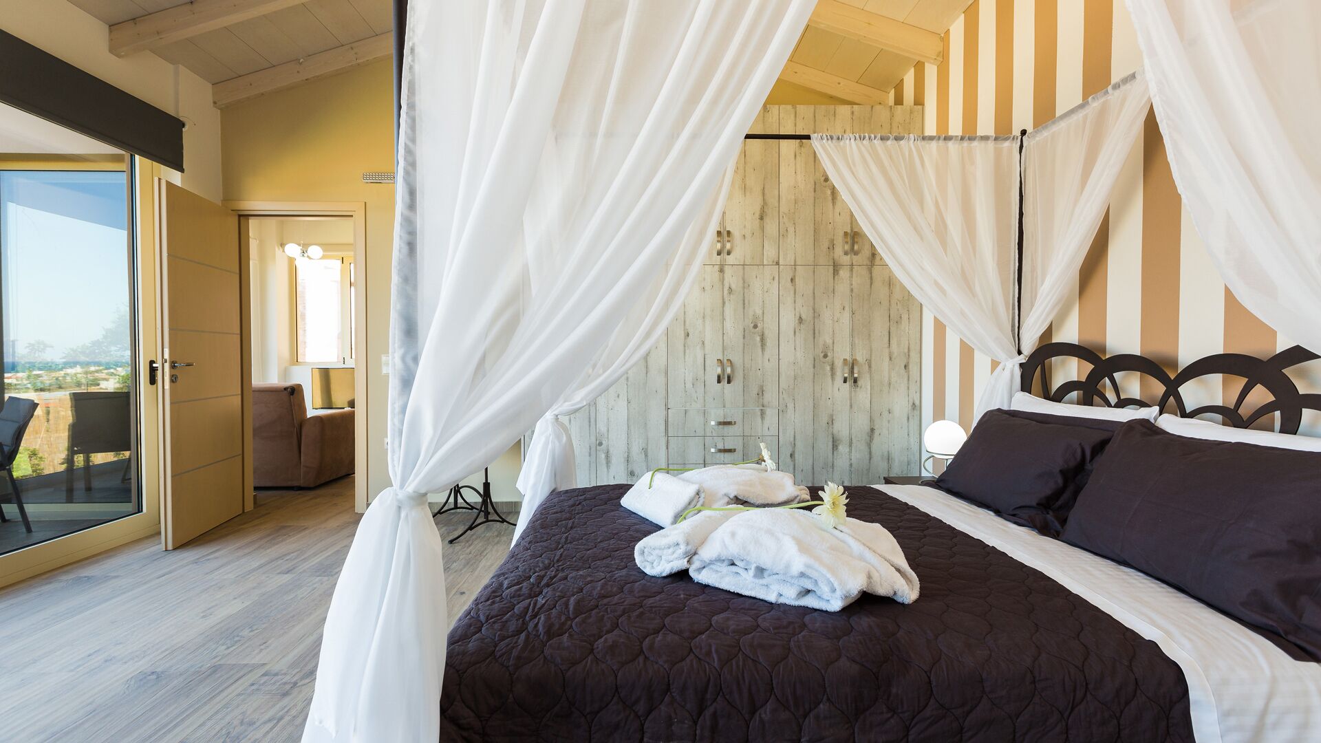 A bedroom with a bed, white curtains, a wooden wardrobe, and a view of the outside.