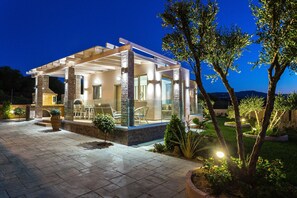A modern house with a covered patio, stone pillars, and a well-lit outdoor area with plants and a lawn. - Villa Kokkinos – Fun & Sun by etouri (Rethymno)