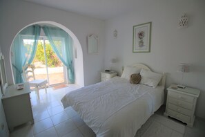 Ein Schlafzimmer mit Bett, Nachttisch, Kommode, Stuhl und einem Fenster mit Vorhängen.