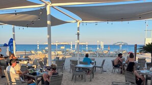 Un espace extérieur aménagé avec des tables et des chaises en osier, donnant sur une plage parsemée de parasols et de chaises longues.