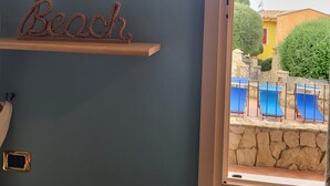 Une salle de bain agrémentée d’une œuvre d’art florale encadrée, d’une étagère sur laquelle est posé un panneau indiquant « Plage », et d’où l’on peut voir, par la fenêtre, des fauteuils bleus installés à l’extérieur.