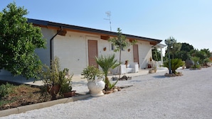 Ein Gebäude mit heller Fassade, einem geneigten Dach und einer Zuwegung mit Kies. Entlang der Auffahrt befinden sich Kübelpflanzen und Gartenmöbel.