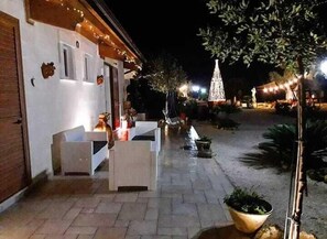 A building with a white exterior, a wooden door, and a small porch area with a bench. There are string lights and a tree with a white top decoration in the background. - B&B Giardino Isabella (Palagiano)