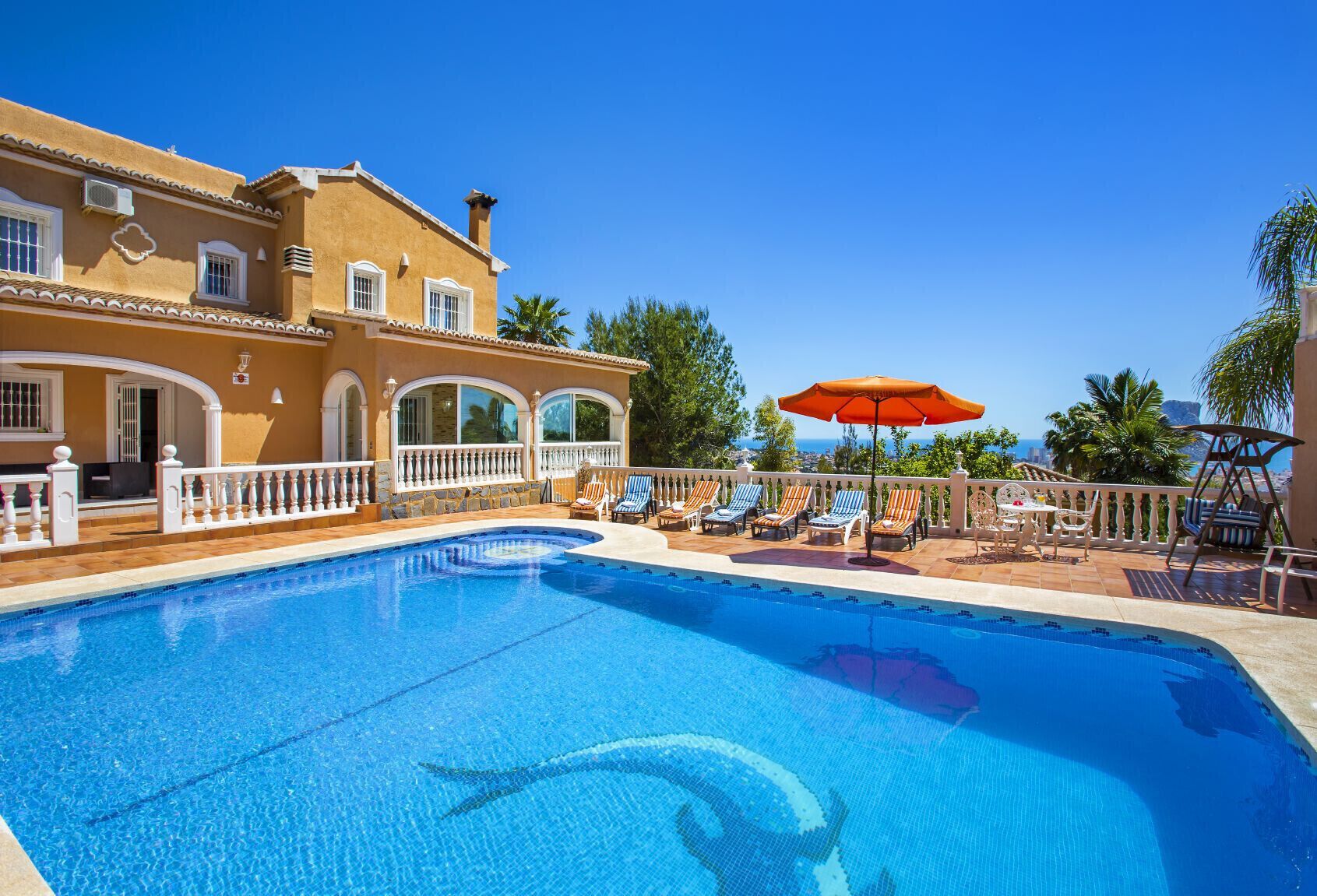 Una piscina con sillas de descanso y sombrillas, una casa grande con ventanas de arco y un cielo azul despejado.