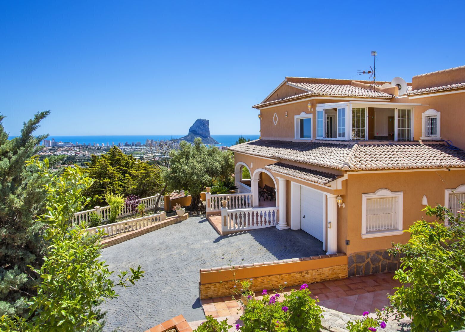 A Mediterranean-style villa with a terrace, a view of the sea, and a prominent rock formation in the background.