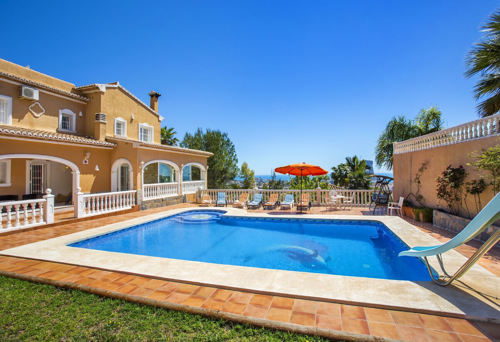 A swimming pool with a slide, surrounded by a white fence and a patio area with lounge chairs and an umbrella.