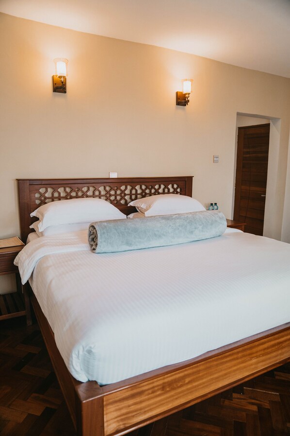 A neatly made bed with white linens and a light blue throw blanket, wooden headboard, two wall-mounted light fixtures, and a wooden door in the background. - Saab Royale Resort (Ilyagaleni)