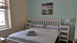 En snyggt bäddad säng med kuddar, ett sängbord med en lampa, en väckarklocka och en tavla på väggen.