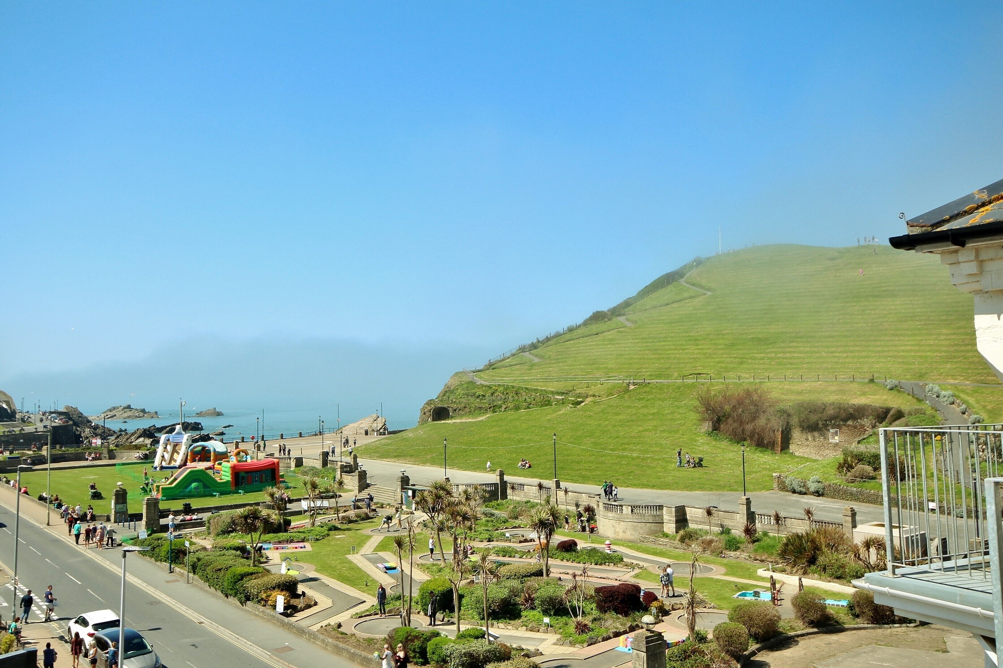 A coastal park with a prominent green hill, a pedestrian path, and a small building with a colorful roof.