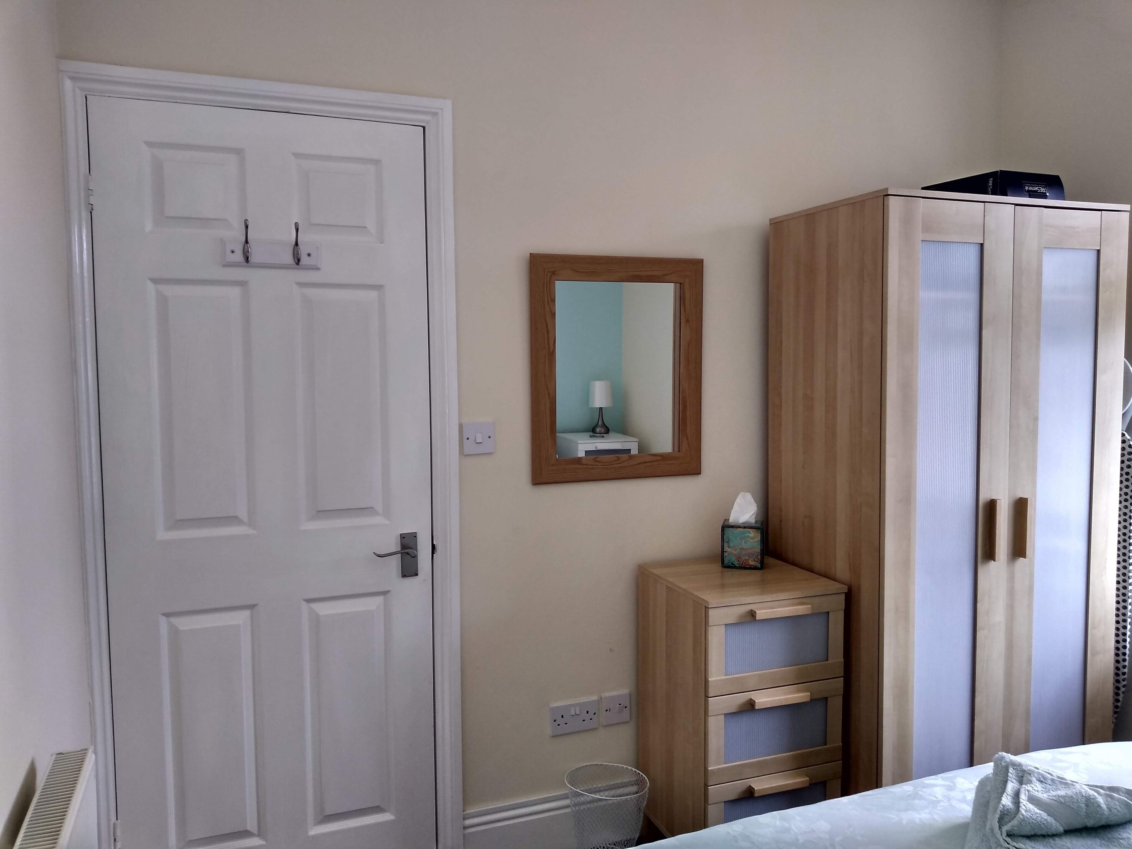 A room with a white door, a wooden cabinet with drawers, a mirror on the wall, and a bed with a white cover.