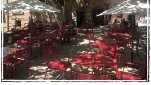 An outdoor cafe with red chairs and tables, stone walls, and umbrellas providing shade. - Village Studio Apartment (Sablet)