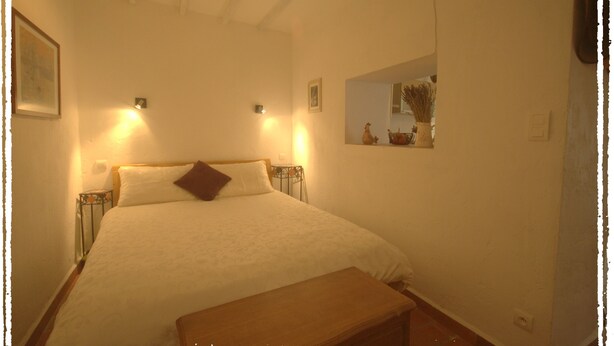 A bedroom with a bed, a bench, a wall-mounted mirror, and two wall sconces. - Village Studio Apartment (Sablet)