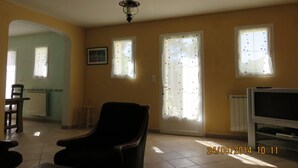 A living room with a black armchair, a television, and a ceiling fan. There are two windows with curtains, a radiator, and a picture on the wall.