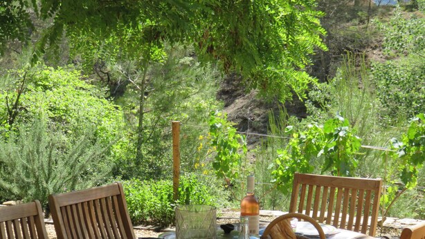 A wooden table set outdoors with chairs, surrounded by lush greenery and a rocky hillside.