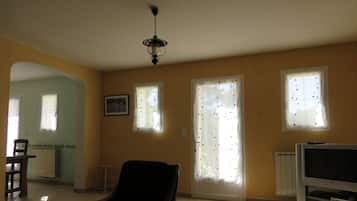 A living room with a black armchair, a television, and a ceiling fan. There are two windows with curtains, a radiator, and a picture on the wall.