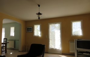 Un salon avec un fauteuil noir, un téléviseur et un ventilateur de plafond. Il y a deux fenêtres avec des rideaux, un radiateur et un tableau accroché au mur.