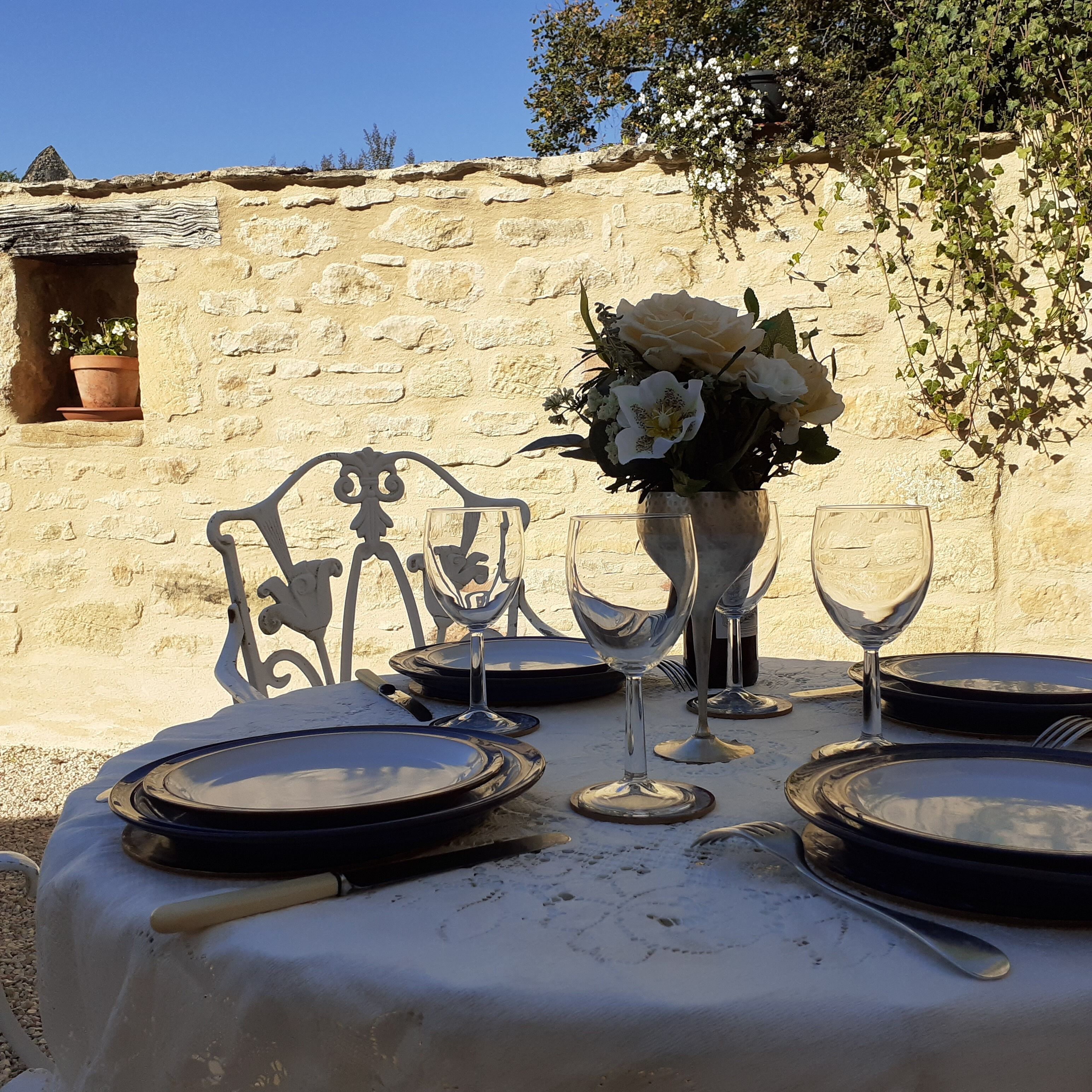 Une table dressée à l’extérieur, avec un mur de pierres en arrière-plan, un vase de fleurs et un ciel dégagé.