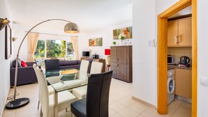 A modern living room with a sofa, dining table, and chairs, a kitchen with wooden cabinets, and a washing machine.