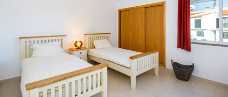 A bedroom with two single beds, a nightstand, a wardrobe, a window with a view, and a laundry basket.