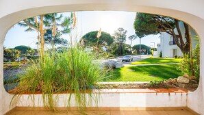 A view through a window with a tiled floor, a grassy area, and a white building with a balcony.